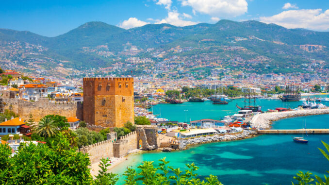 Haartransplantation für Frauen Panoramablick auf den Roten Turm und den Hafen von Alanya mit türkisblauem Meer und Bergen im Hintergrund.
