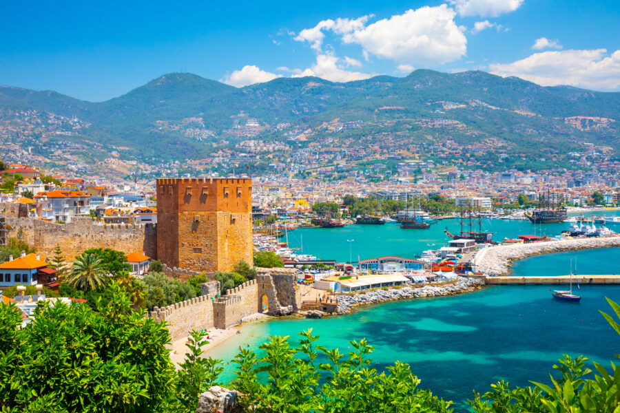 Panoramablick auf den Roten Turm und den Hafen von Alanya mit türkisblauem Meer und Bergen im Hintergrund.